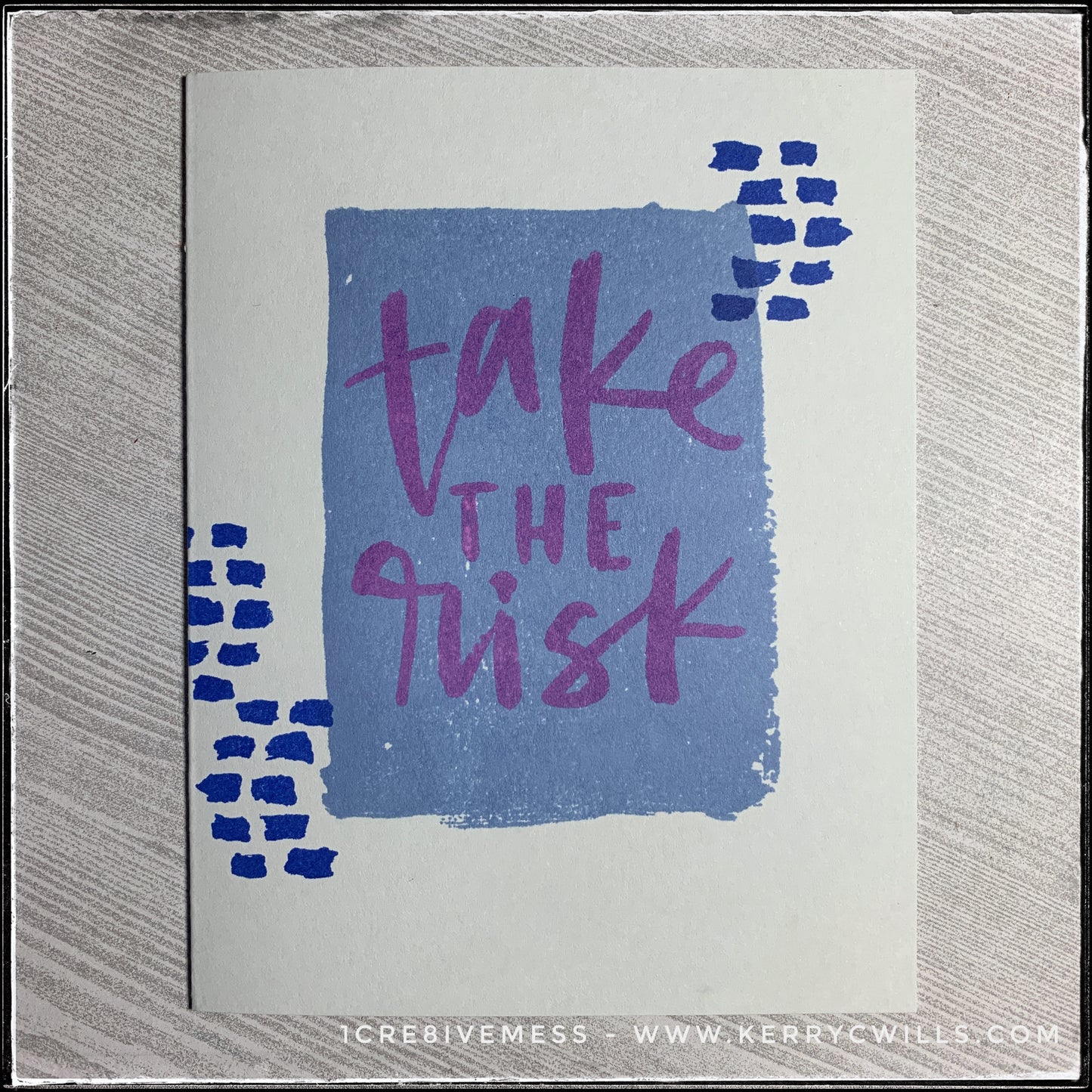 A flat-lay view of this encouraging handmade card, the message stamped front and center reads "take the risk" in purple ink. The sentiment is layered over a blue rectangle on the middle of the soft grey card base. Small dark blue dashed lines surround the sentiment on either side.