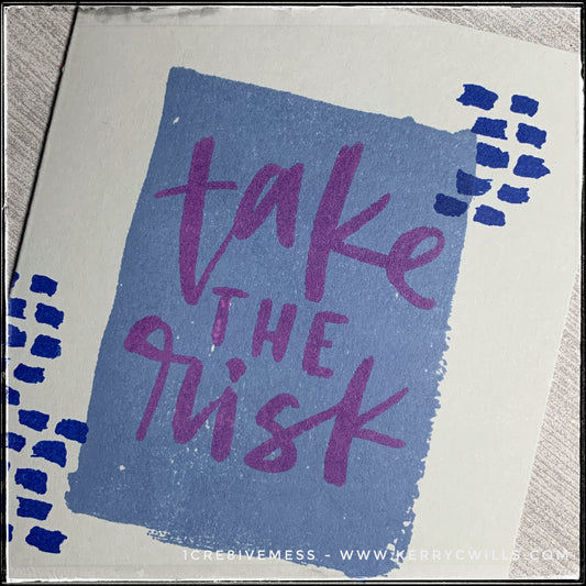 An up-close, angled view of the main elements on the front of this handmade card. A blue rectangle that appears to have a watercolored finish creates a nice bold background for the stamped sentiment "take the risk." Smaller, darker blue dashed lines accent the sentiment on either side.