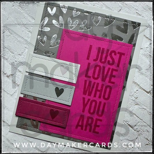 Handmade card from Day Maker Cards, stamped with 'I Just Love Who You Are' text on a pink background with heart designs, displayed against a silver foiled background.