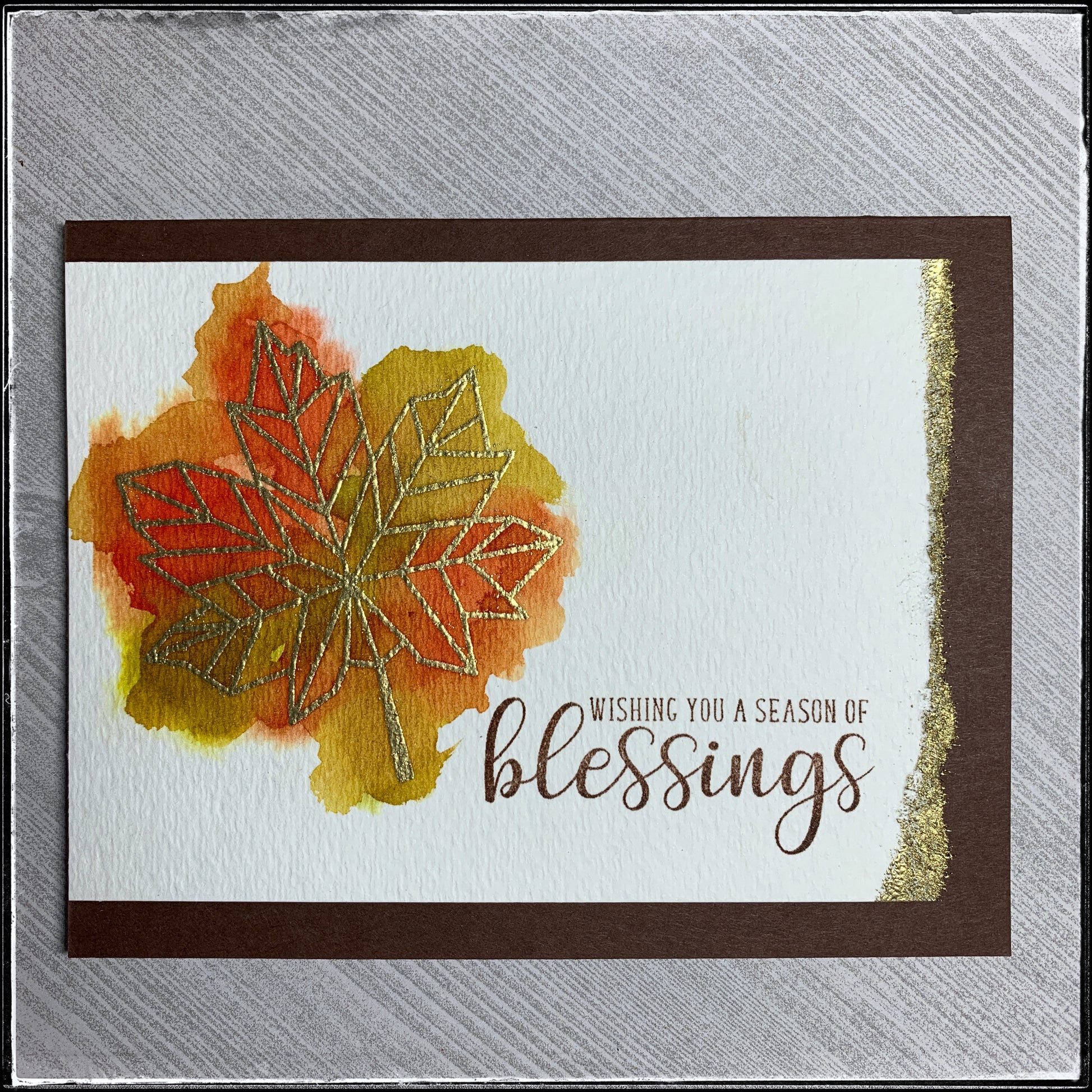 A handmade card from 1cre8ivemess with a watercolored background behind a heat embossed geometric gold leaf. Stamped with the sentiment "Wishing you a season of blessings" in brown ink. 
