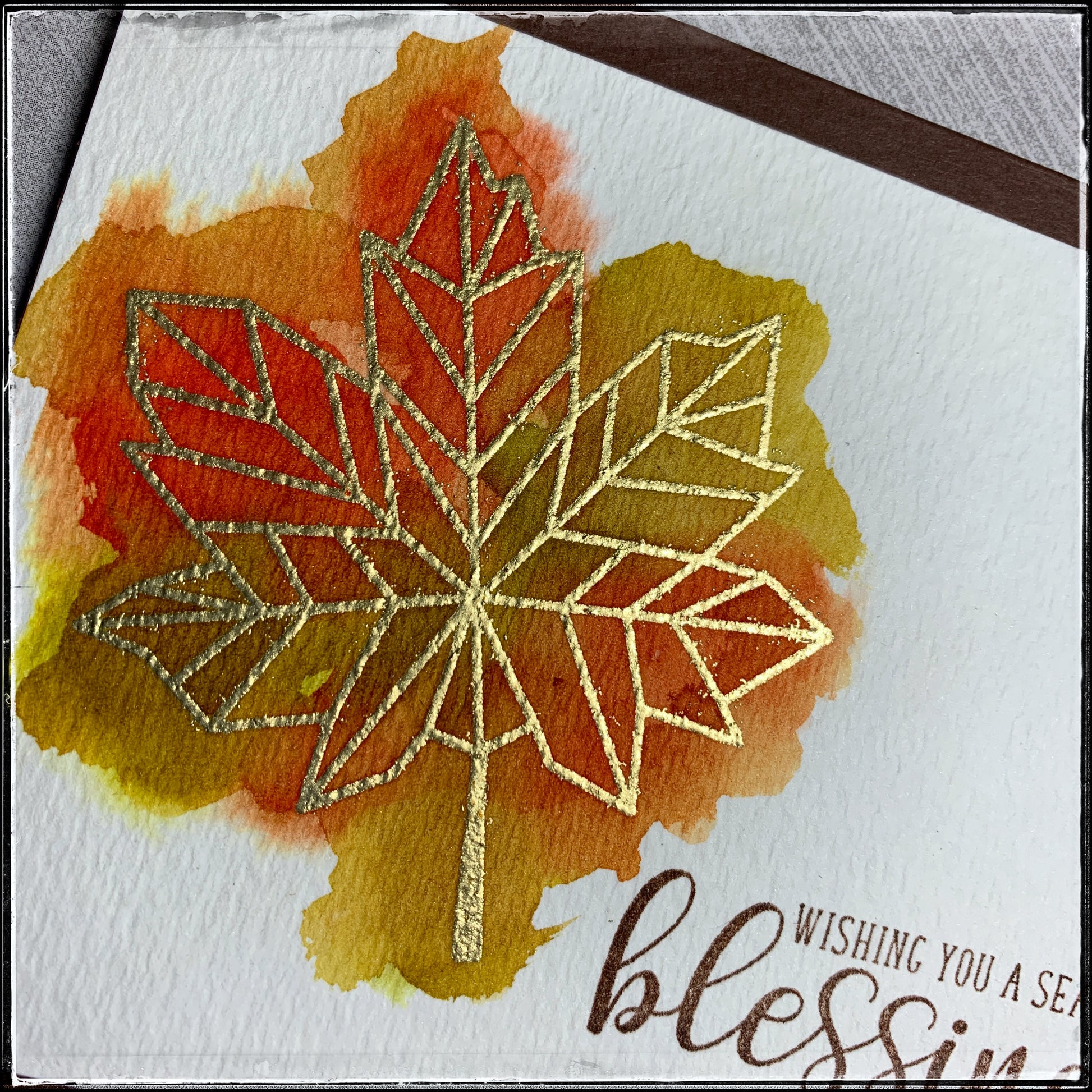 Up close details of a handmade card from 1cre8ivemess with a watercolored background behind a heat embossed geometric gold leaf. Stamped with the sentiment "Wishing you a season of blessings" in brown ink. 