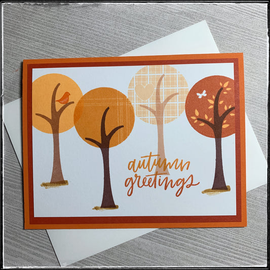 A fall-themed greeting is the main sentiment on this handmade card! "Autumn greetings" is stamped in an ombre color scheme with orange fading from light to dark alongside four trees. Each tree top is a circle in a shade of orange as the concept of this card is a monochromatic color scheme. Each tree top is slightly different, both in pattern and in color. They are staggered across the white card panel which is layered on a darker orange panel which is layered on the classic orange card base. A white envelop