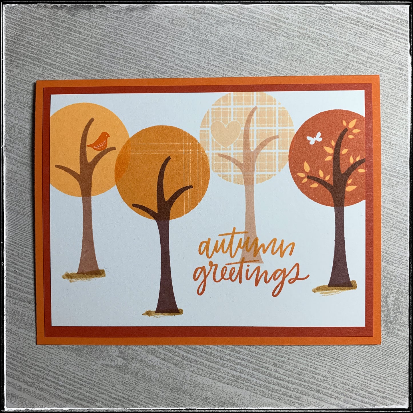 The flat-lay view of this seasonal handmade card. "Autumn greetings" is stamped in shades of orange and overlaps the trunk of one of the four trees that are stamped across the front panel. Each tree top is a slightly different shade of orange, creating a mostly monochromatic look on this handmade card. 