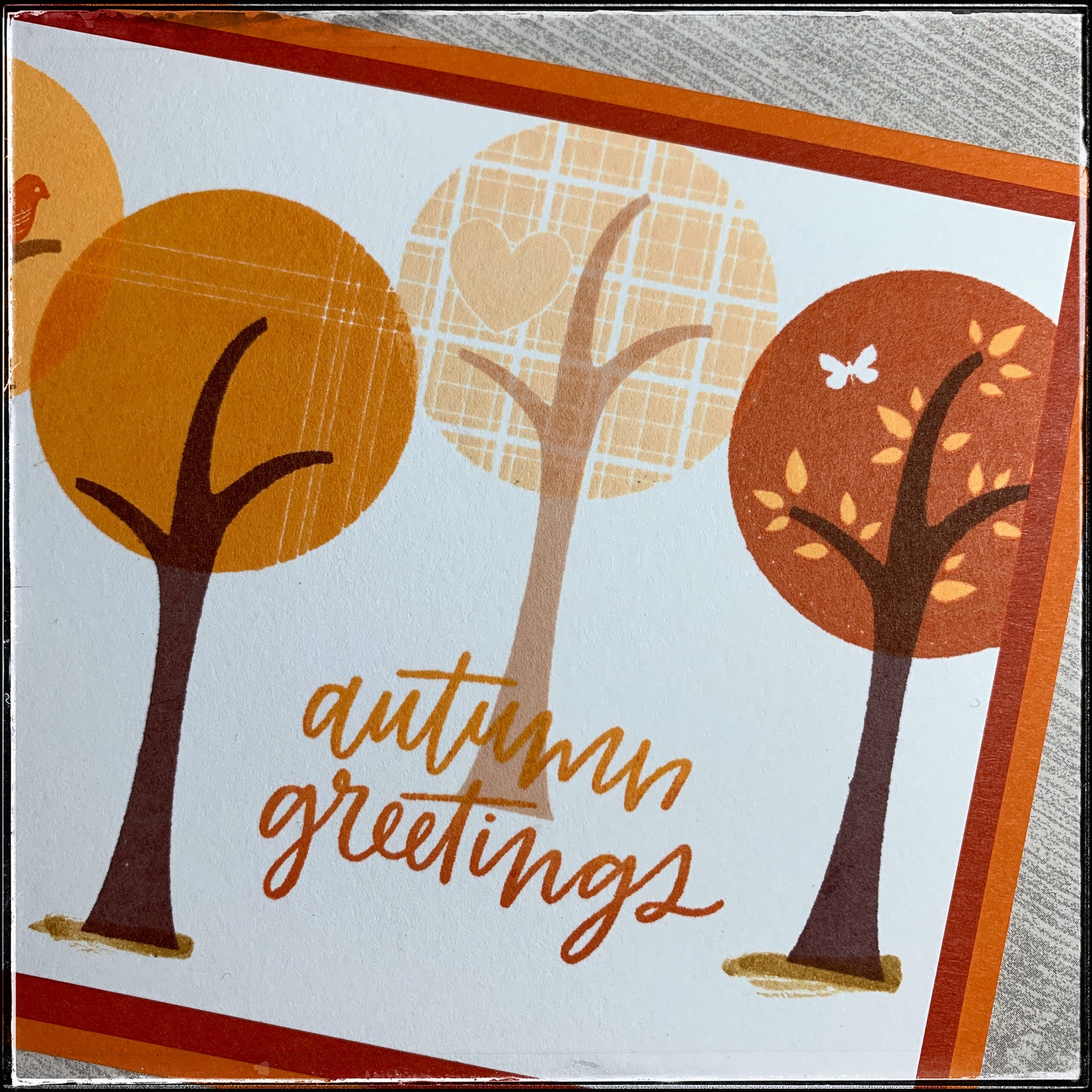An up close detailed view of several of the stamped trees and the details on the tree tops. Thin striped lines are crisp and clear in a medium shade of orange. The lightest colored tree top is a beautiful plaid pattern with an inlaid heart design. The third tree top shown is the darkest color orange and features some lighter orange colored leaves. The sentiment "autumn greetings" overlaps the trunk of the middle tree in two shades of orange ink which are blended in an ombre color scheme. 
