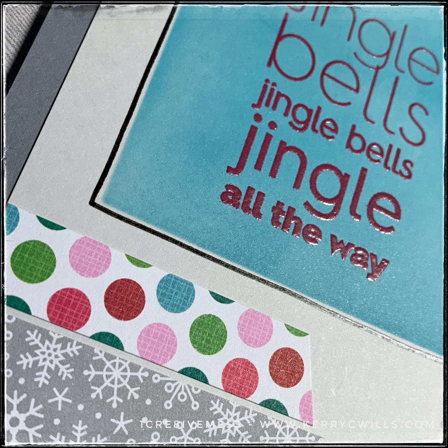 An angled flat-lay view of the embossed sentiment [Jingle bells jingle bells jingle all the way] in red ink and clear powder. There's a gorgeous sheen on this card front, but it doesn't take away from the design. Two banners with angled fish-tail ends accent the light grey card panel below the sentiment. They feature polka dots in reds, pinks, greens, and aquas as well as tiny snowflakes. 