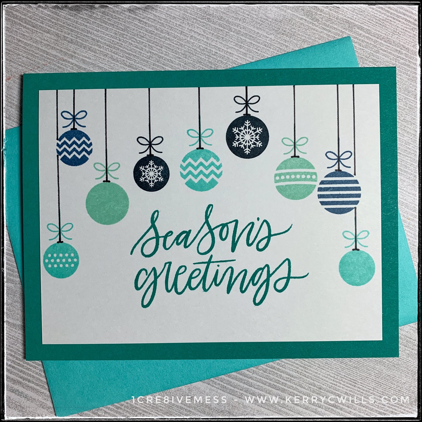 This handmade holiday card features the main sentiment "Season's greetings" which is stamped in a dark turquoise color, coordinating with the card base. A variety of patterned ornaments hang from the top of the panel, surrounding the sentiment. The ornaments are stamped in shades of aqua, turquoise and navy blues, while hand drawn black strings reach to the top of the card panel as if they are really hanging. A turquoise envelope is included and layered beneath the card. 
