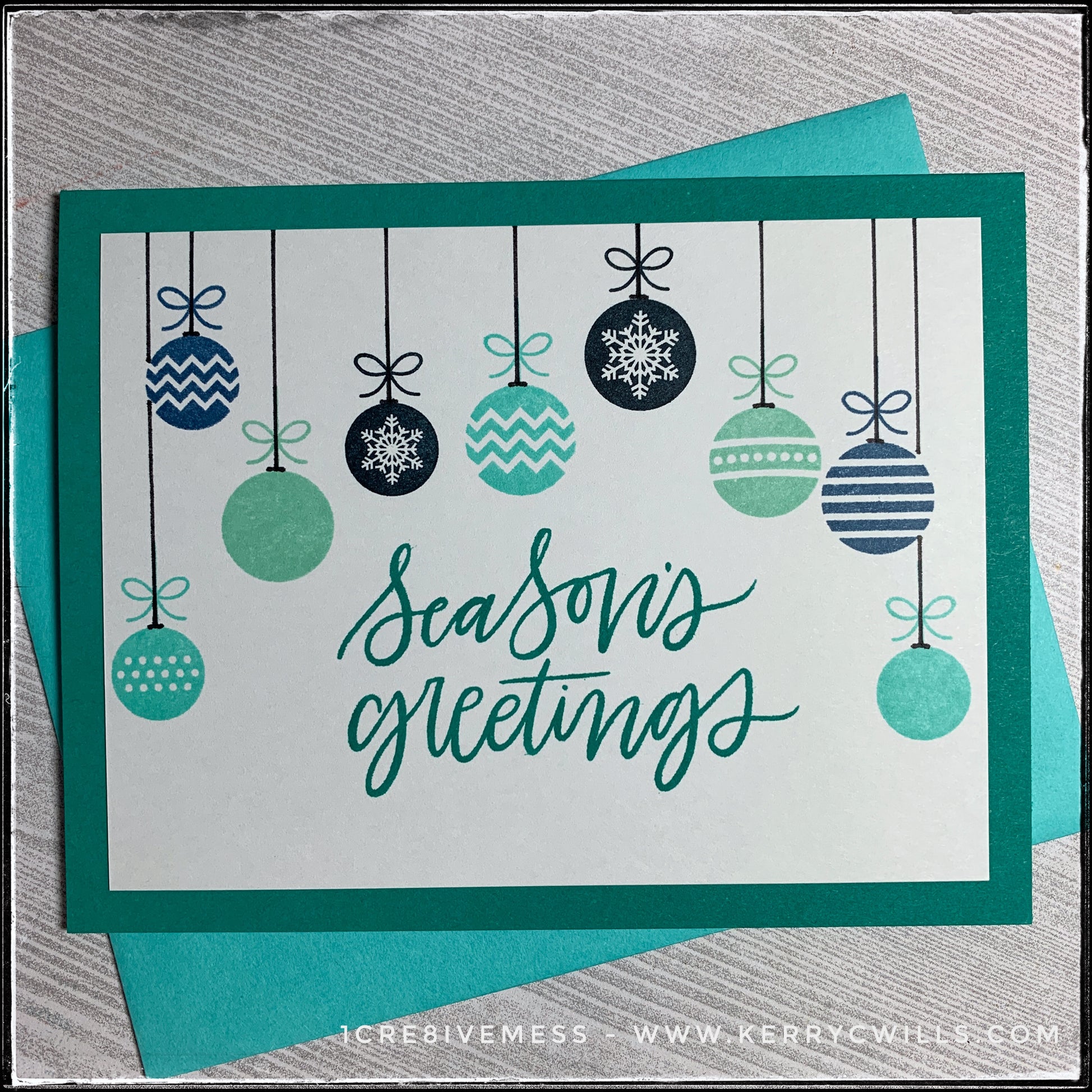 This handmade holiday card features the main sentiment "Season's greetings" which is stamped in a dark turquoise color, coordinating with the card base. A variety of patterned ornaments hang from the top of the panel, surrounding the sentiment. The ornaments are stamped in shades of aqua, turquoise and navy blues, while hand drawn black strings reach to the top of the card panel as if they are really hanging. A turquoise envelope is included and layered beneath the card. 