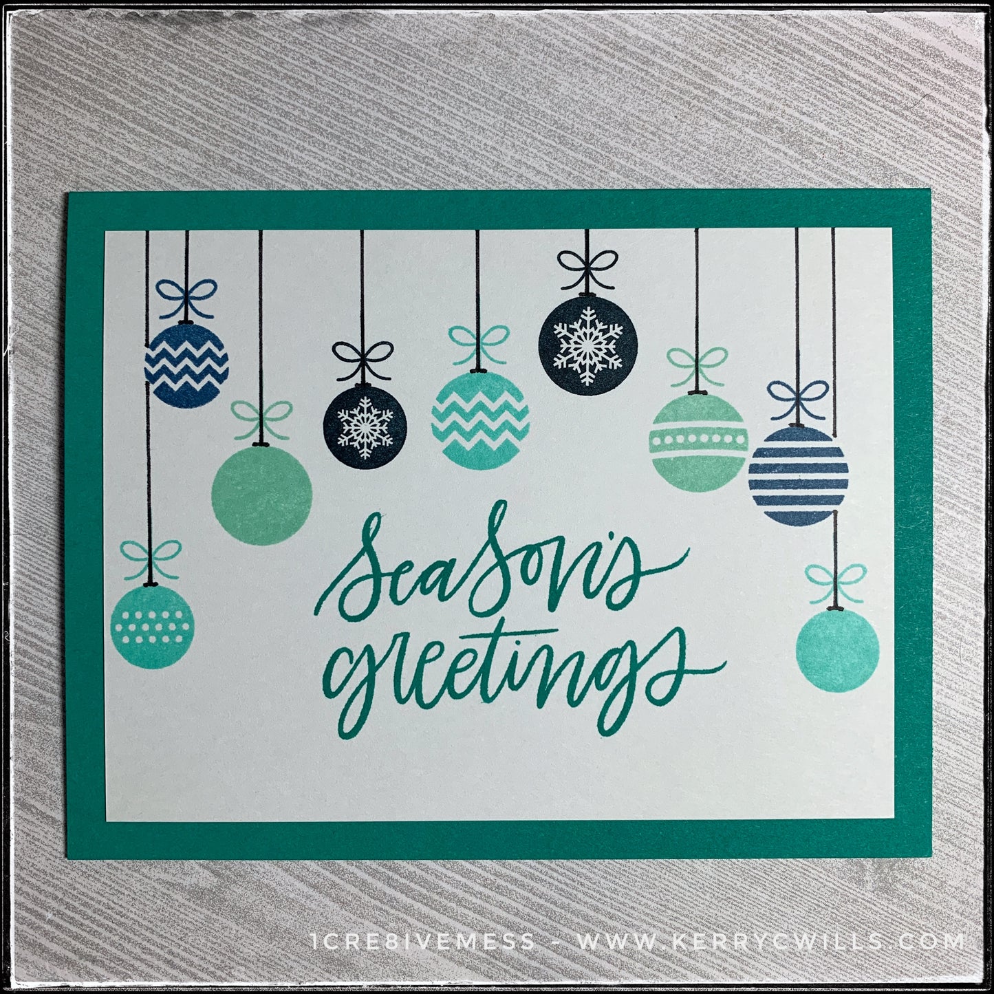 A classic flat-lay of this handmade holiday card, the sentiment "season's greetings" is stamped in the center of the card front. The color coordinates with the color of the card base. A series of nine hanging ornaments graces the top portion of the card, cascading along the top. Shades of aquas, turquoises and deep blues evoke a wintery feeling. 