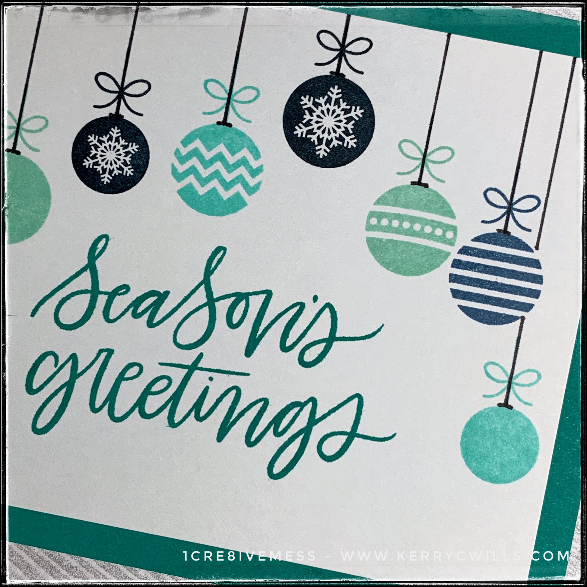 An up-close, angled view of the detail on several of the stamped ornaments. A hand drawn black line extends from the ornament to the top of the card panel, as if they are hanging. Patterns of the ornaments include chevrons, polka dots, solid balls, stripes and snowflakes. The sentiment "season's greetings" is nice and big and bold in the center of this handmade card. 