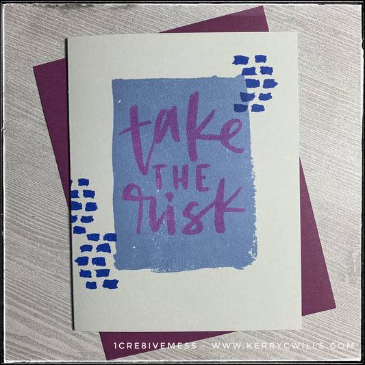 This handmade card is of the encouragement type, with the big bold message "take the risk" stamped in purple ink over a watercolor-esque rectangle on the light grey card front. Small darker blue dashed lines accent the sides of the rectangle. A deep purple envelope is included and is shown layered under the card. 