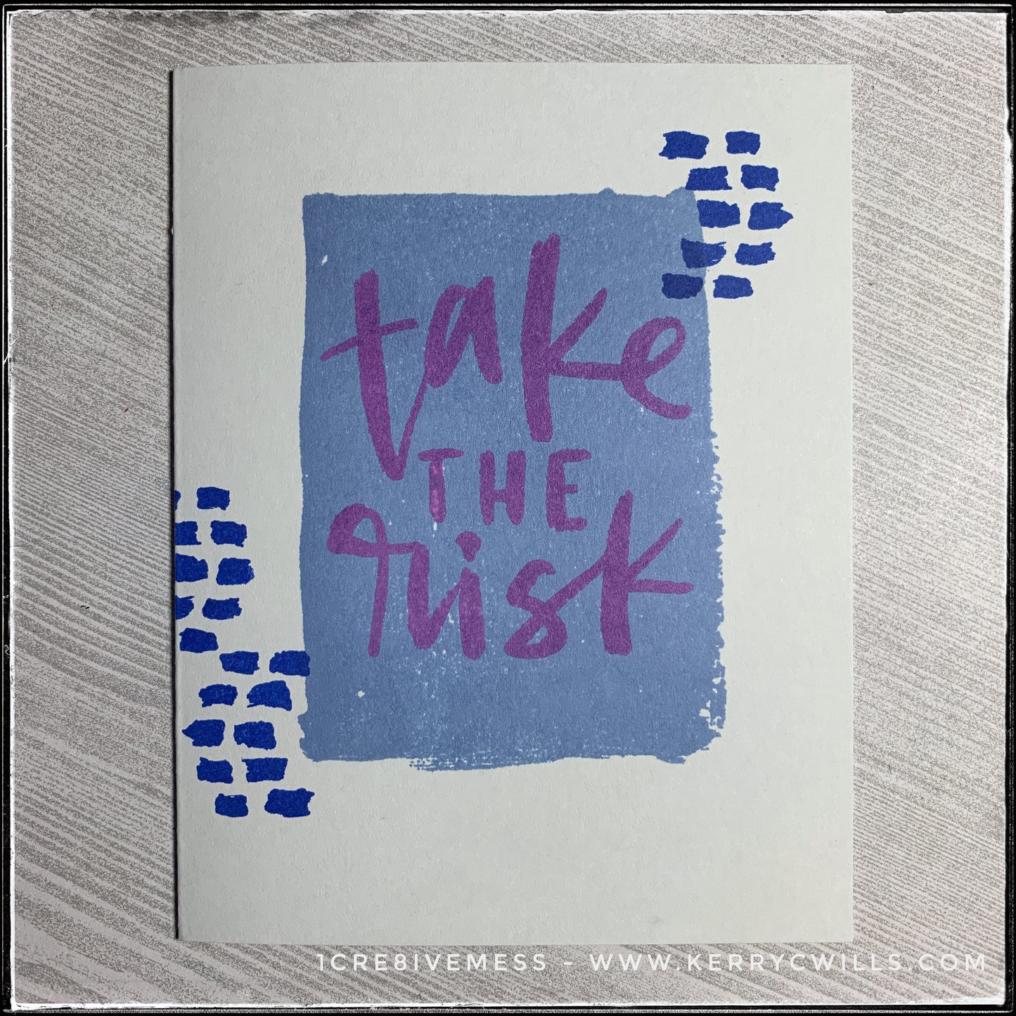 A flat-lay view of this encouraging handmade card, the message stamped front and center reads "take the risk" in purple ink. The sentiment is layered over a blue rectangle on the middle of the soft grey card base. Small dark blue dashed lines surround the sentiment on either side. 