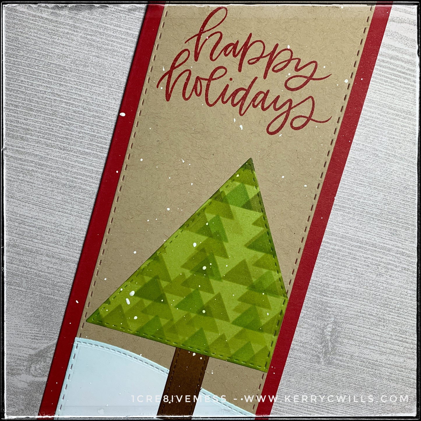 This angled flat-lay view includes the sentiment "happy holidays" in red ink, in a scripty font. The red color coordinates with the color of the slimline card base. The faux-stitched detail is visible around the edges of the card panel, the tree, tree trunk and snowbank. Inked patterns add dimension and detail. 