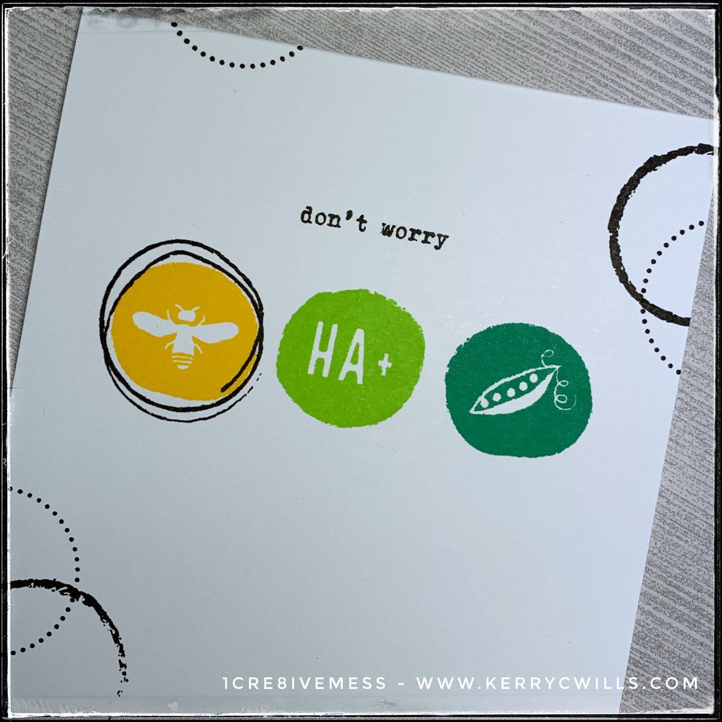 An angled view of the front panel on this handmade card features three colorful circles in shades of yellow, lime green and grass green. On each of the circles is an image or a series of letters that combine to form a sentiment, along with the stamped words above them. When reading the entire card front, it reads "don't worry be happy" The black and white classic design is timeless and this card is sure to make the recipient smile. 
